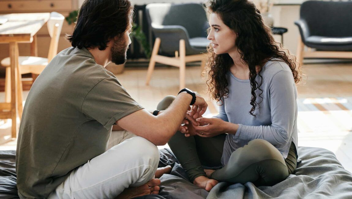 Una pareja sentada en la cama habla con las manos agarradas. Ella tiene gesto de preocupación. Él no muestra el rostro pero por la forma en la que da la mano parece estar preocupado.